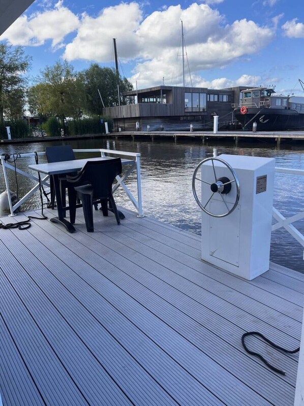 A wooden deck with a table and chairs, a white electrical box, and a boat docked in the background.