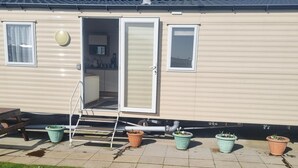 A mobile home with a kitchen area visible through an open door, a small window, and a chimney. There are steps leading to the door and several potted plants outside.