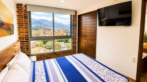 A bedroom with a bed, a TV mounted on the wall, a window with a city view, and a brick wall.