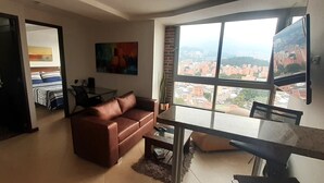 Ein Wohnzimmer mit braunem Ledersofa, einem Glastisch und einem Esstisch mit Stühlen. Ein großes Fenster bietet Blick auf die Stadt.