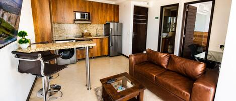A modern living room with a leather sofa, a coffee table, and a kitchen area with wooden cabinets, a refrigerator, and a washing machine.