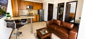 A modern living room with a leather sofa, a coffee table, and a kitchen area with wooden cabinets, a refrigerator, and a washing machine.