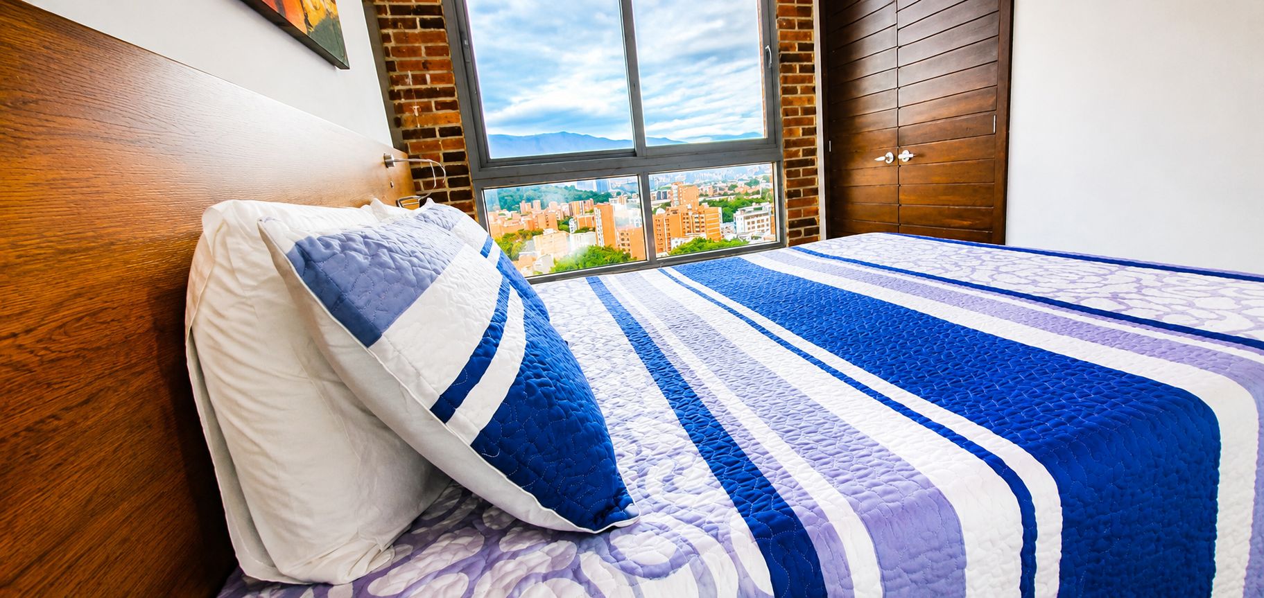 A bedroom with a bed, a window with a city view, and a wooden headboard.