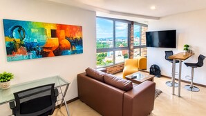 A modern living room with a brown leather sofa, a glass coffee table, a TV mounted on the wall, and a large window offering a city view. - Laureles Penthouse rooftop Suite (Medellín)