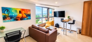 Una sala de estar moderna con un sofá de cuero marrón, una mesa de centro de vidrio, un televisor montado en la pared y un amplio ventanal con vistas a la ciudad.