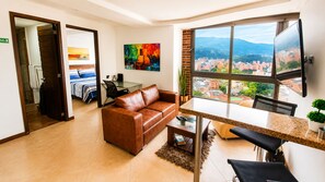 A modern living room with a brown leather sofa, a dining table with chairs, a large window with a city view, and a painting on the wall. - Laureles Penthouse rooftop Suite (Medellín)