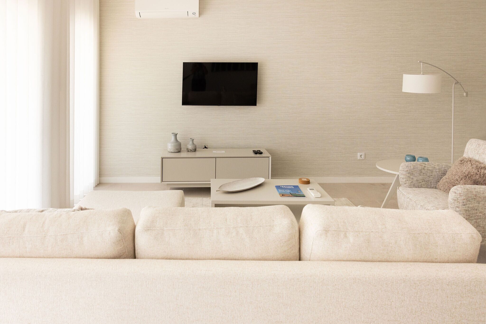 A modern living room with a sofa, a TV mounted on the wall, a side table with a remote control, and a lamp.