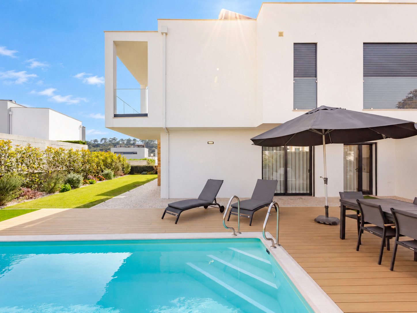 Un área de piscina moderna al aire libre con sillas de descanso y una mesa, rodeada por una terraza de madera y un edificio blanco con grandes ventanales.
