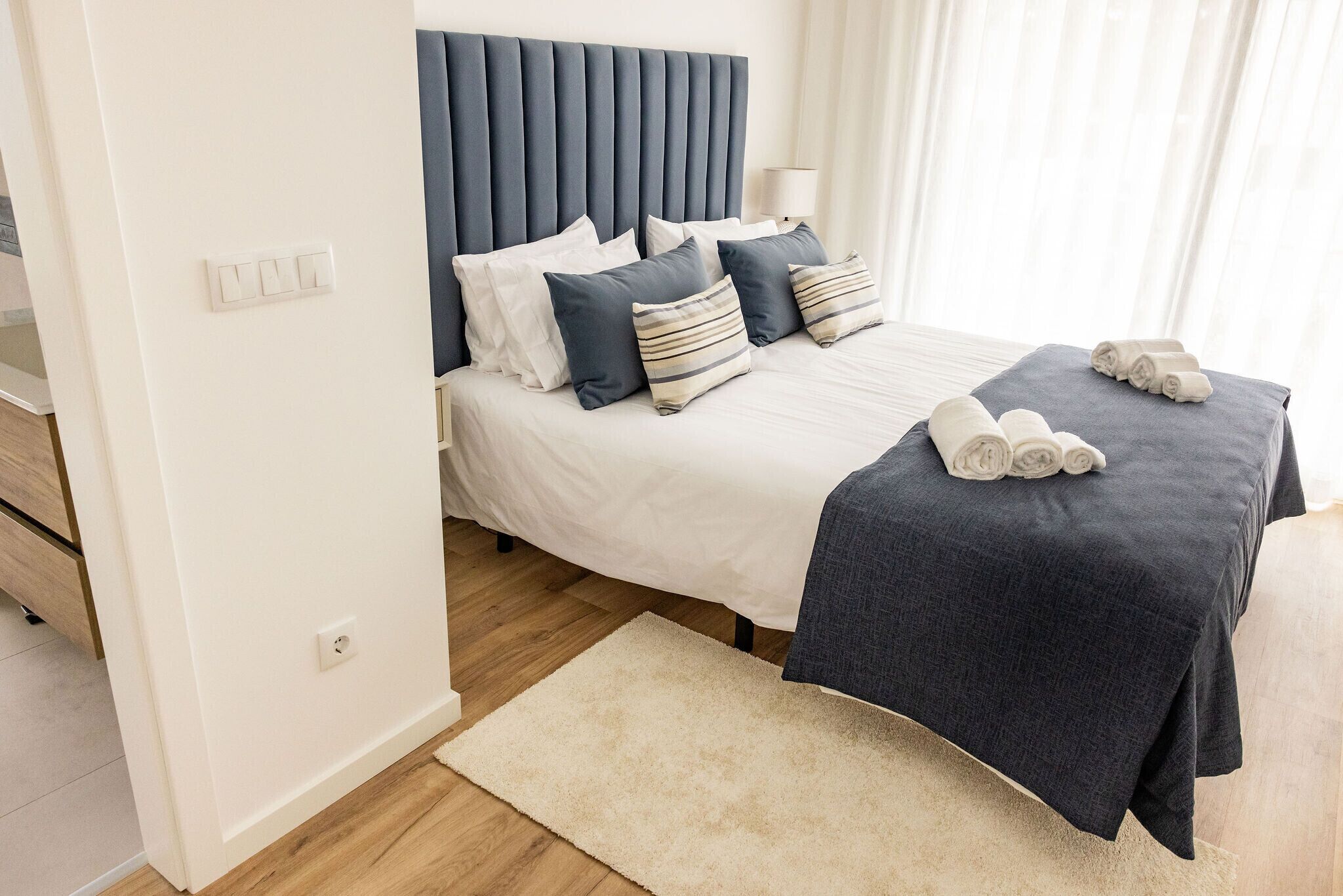A neatly made bed with a dark headboard, white bedding, and a few pillows. There is a rolled towel on the bed and a white rug on the floor.