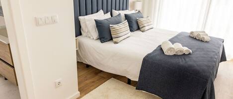 A neatly made bed with a dark headboard, white bedding, and a few pillows. There is a rolled towel on the bed and a white rug on the floor.