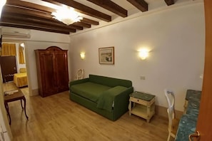 Una habitación con piso de madera, un sofá verde, un armario de madera, una mesita pequeña y una lámpara fijada a la pared.