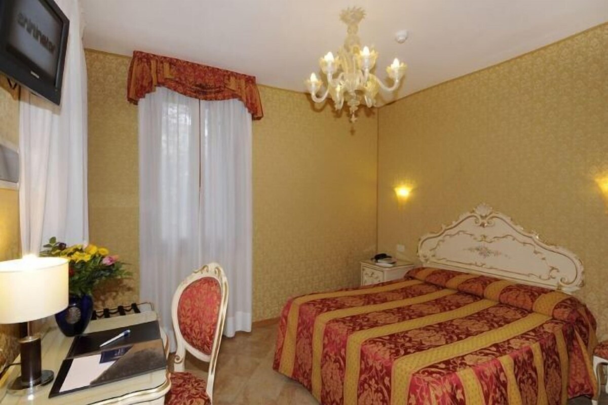 Una habitación de hotel con cama, escritorio con lámpara, silla, televisor y candelabro.