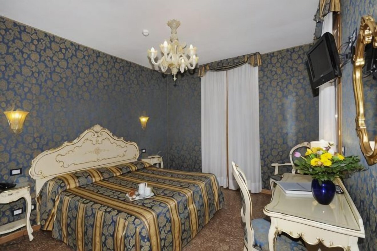 A hotel room with a double bed, a desk with a chair, a television, and a vase with flowers.