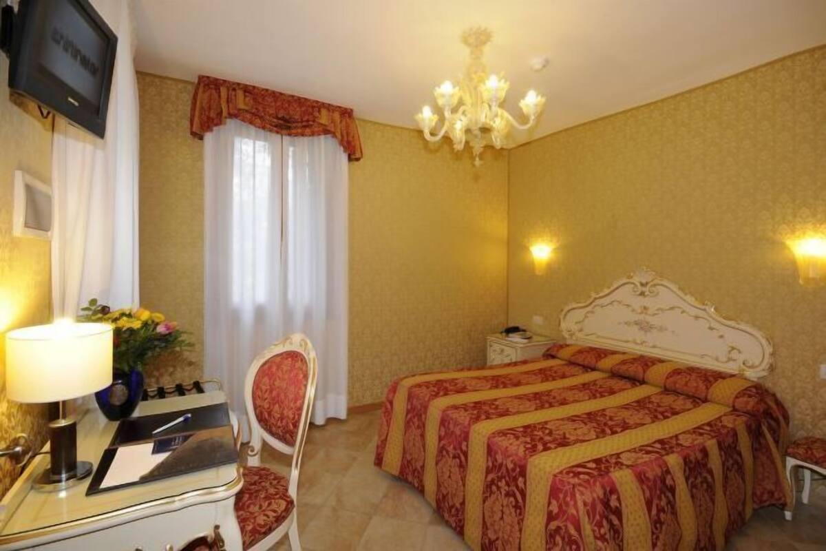 Habitación de hotel con cama, escritorio con lámpara, televisión, silla y candelabro.
