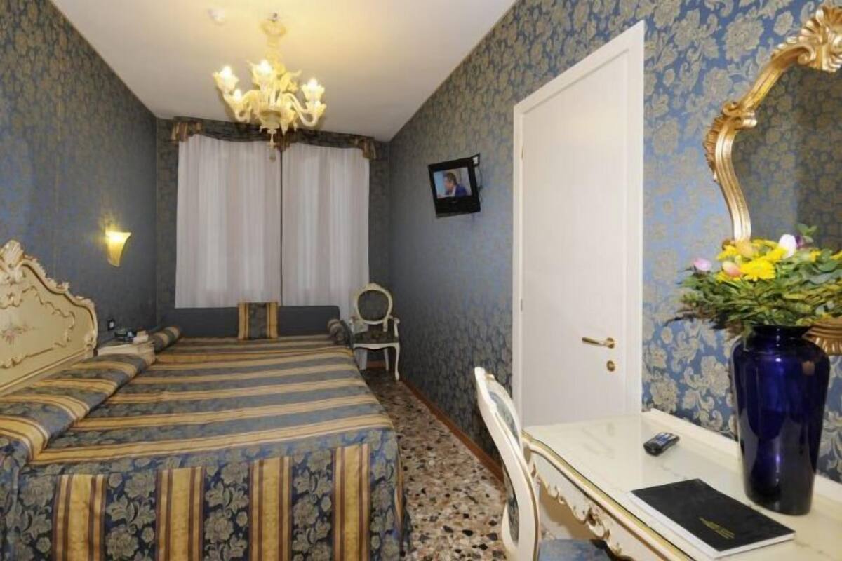 Una habitación con cama, escritorio, silla, espejo, candelabro y un florero con flores.