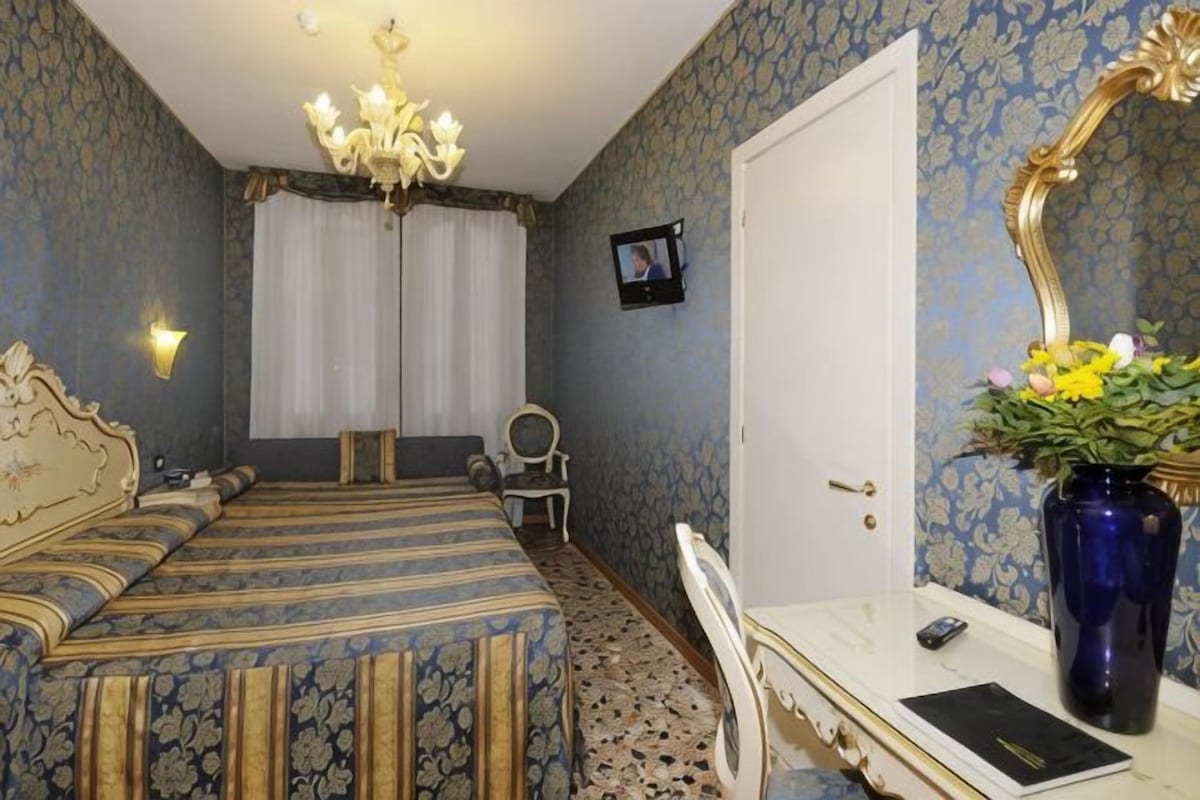 A bedroom with a bed, a desk, a chair, a dresser, a mirror, a chandelier, and a door.