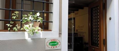A building entrance with a potted plant on the ledge, a door with a decorative grille, and a sign with a logo.