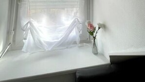Une pièce avec une fenêtre recouverte d’un rideau léger, un vase contenant une seule fleur et un mur blanc.