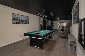 A pool table in a room with a dark ceiling, a painting on the wall, and a chair.