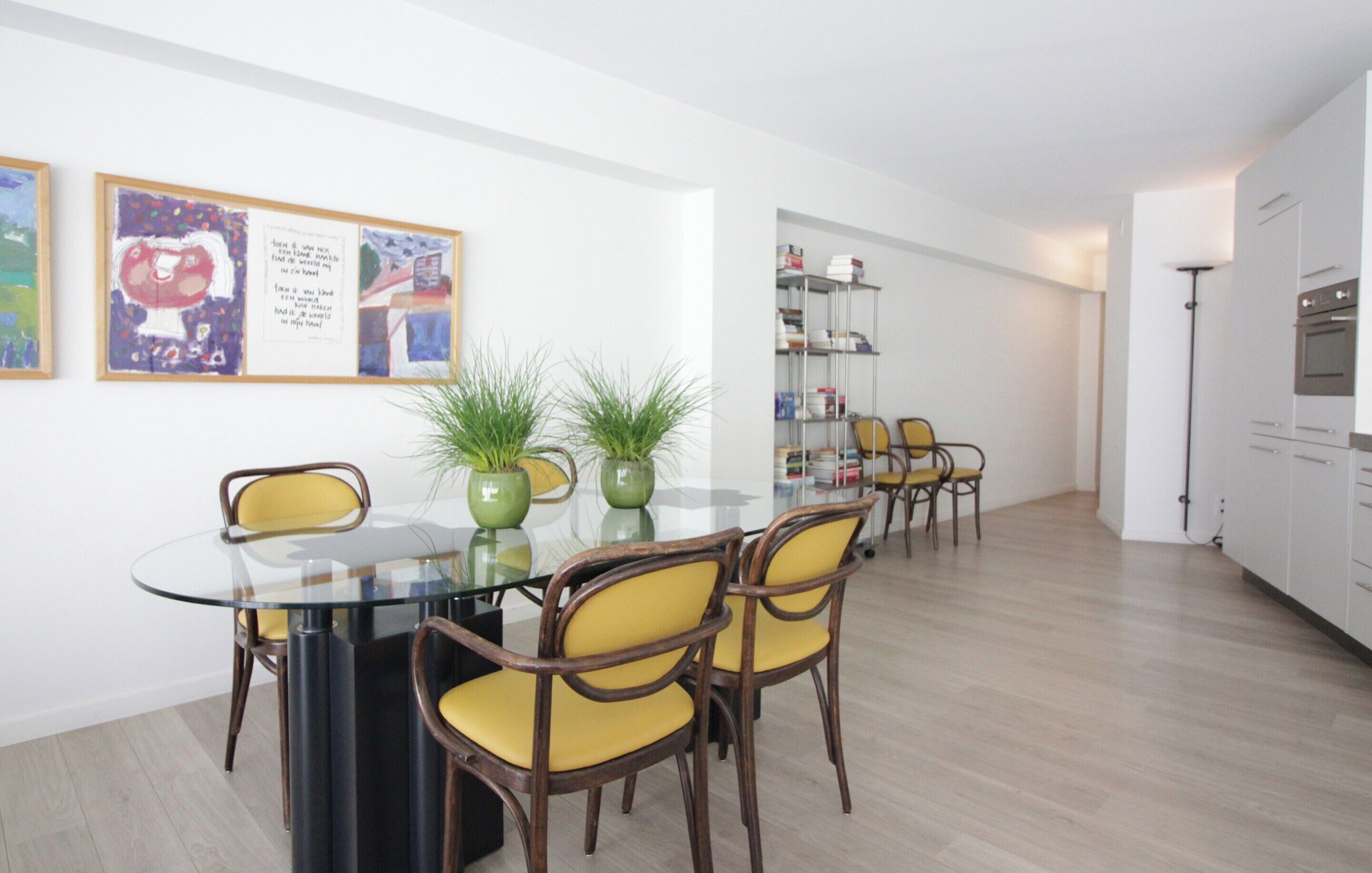 A modern dining area with a glass table and wooden chairs, surrounded by white walls and a wooden floor.