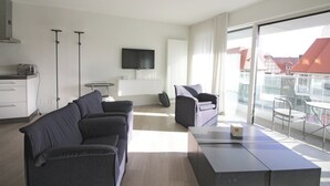 A modern hotel room with a sofa, a small table, a flat-screen TV, and a balcony with a view of buildings. - Apartment in Knokke Near Driehoeksplein (Knokke-Heist)