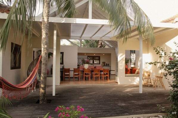 Un espacio al aire libre techado con una terraza de madera, una hamaca y una zona de comedor con sillas y una mesa.
