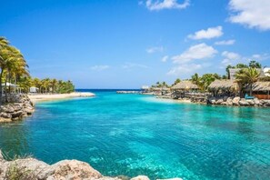 Un mar turquesa transparente con una playa arenosa, palmeras y cabañas de techo de paja.