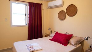 Un dormitorio con una cama, una ventana con cortinas rojas, aire acondicionado y dos tapices de pared.
