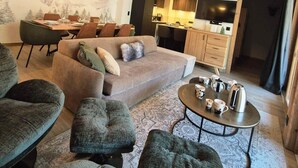 A modern living room with a sofa, ottomans, and a coffee table. A dining area with chairs and a table is visible in the background.