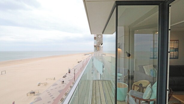 A balcony with a view of a beach and a glass door leading to a room with a sofa and a chair.