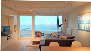 A modern living room with a sofa, a dining table, and a TV. There is a large window with a view of the sea.