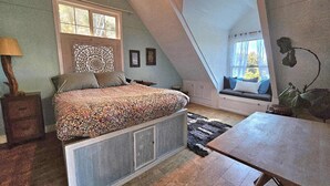 Un dormitorio con techo inclinado, una cama con ropa de cama estampada, un banco junto a la ventana con cojines y una mesa.