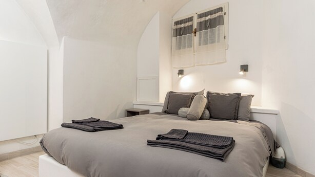 A bedroom with a large bed, two bedside tables, a nightstand, a window with a lace curtain, and a wall-mounted light fixture.