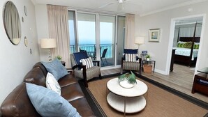 A modern hotel room with a brown leather sofa, two armchairs, a round coffee table, and a balcony with a view of the ocean. - Emerald Beach 1130 (Panama City Beach)