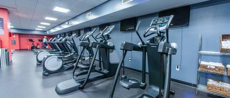 A gym with multiple treadmills and elliptical machines, a red wall, and a shelf with baskets.