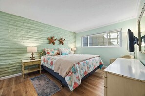 A bedroom with a brick-patterned wall, a bed with floral bedding, a nightstand with a lamp, and a window with blinds. - Rio del Mar Paradise with sweeping oceanviews! (Aptos)