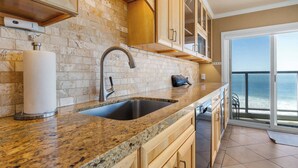 A kitchen with a granite countertop, a stainless steel sink, wooden cabinets, and a view of the ocean through a sliding glass door.