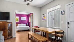 A room with a wooden dining table, a bench, a bed with white bedding, a TV, and a window with blinds.