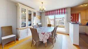 A dining area with a round table, chairs, a cabinet with glass doors, a chandelier, and a view through a window with striped curtains. - Duplex in Knokke Near North Sea Beach (Knokke-Heist)