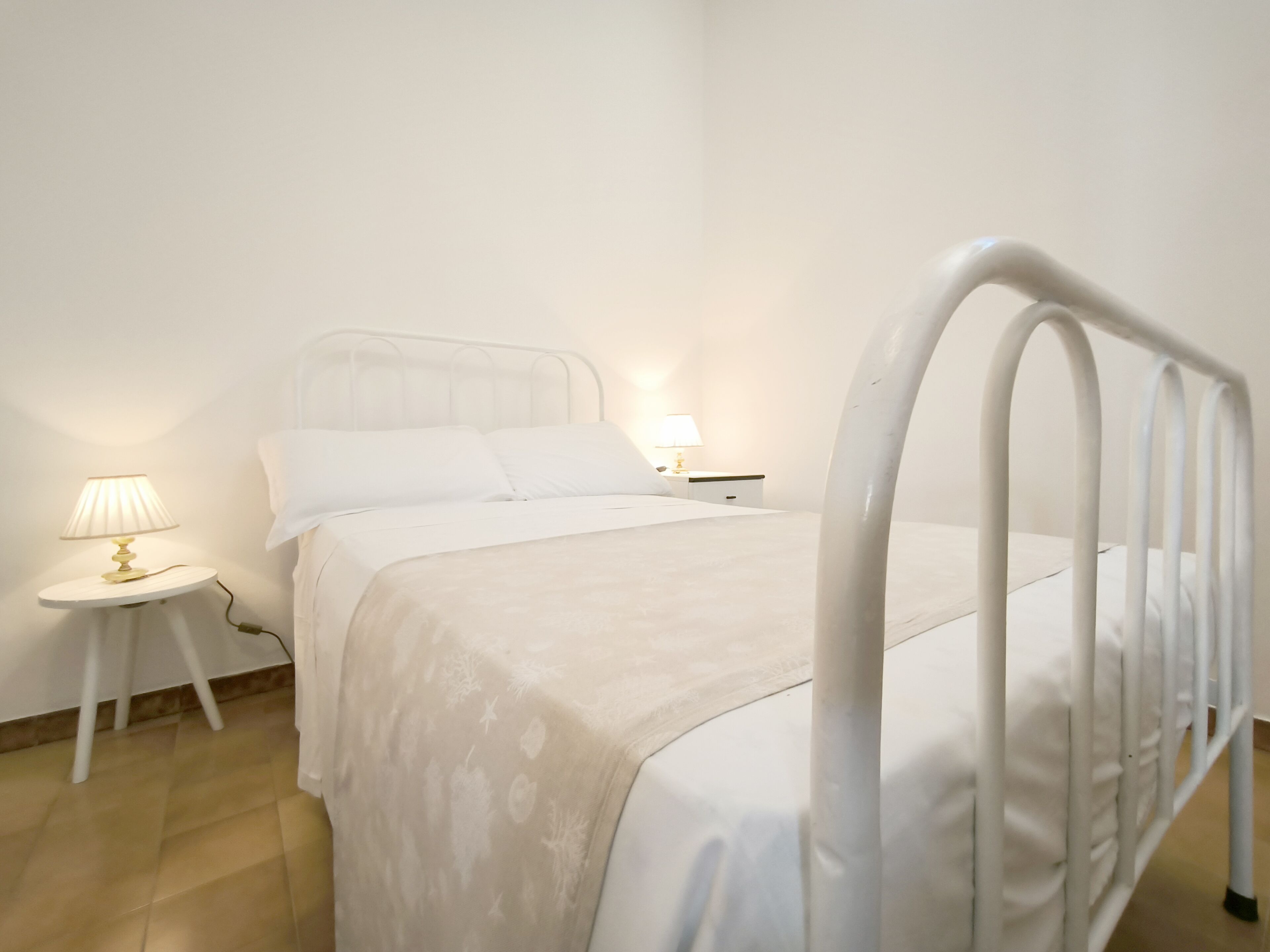 Ein schlicht eingerichtetes Schlafzimmer mit einem weißen Metallbettgestell, einem runden Nachttisch mit Lampe und einem Beistelltisch ebenfalls mit Lampe.