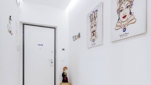 Ein Flur mit einer weißen Tür, weißen Wänden und einem hellen Holzboden. An der Wand hängen zwei gerahmte Kunstwerke und auf dem Boden steht ein kleines dekoratives Objekt.