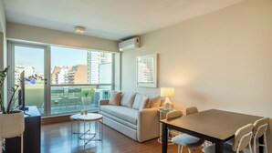 Una sala de estar moderna con un sofá, una mesa de comedor y sillas. Se aprecia una ventana grande que ofrece vistas a la ciudad.