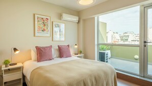 Un dormitorio moderno con una cama grande, dos mesitas de noche, una mesita auxiliar, una ventana con vista a la ciudad y dos unidades de aire acondicionado fijadas a la pared.