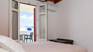 Une chambre avec un lit, une petite table et une chaise. Une porte ouverte donne sur un balcon où se trouvent une table et des chaises.