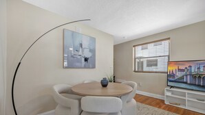 Una sala moderna con una mesa redonda de madera, dos sillones grises, una lámpara de pie, un cuadro, un televisor y una ventana con persianas.