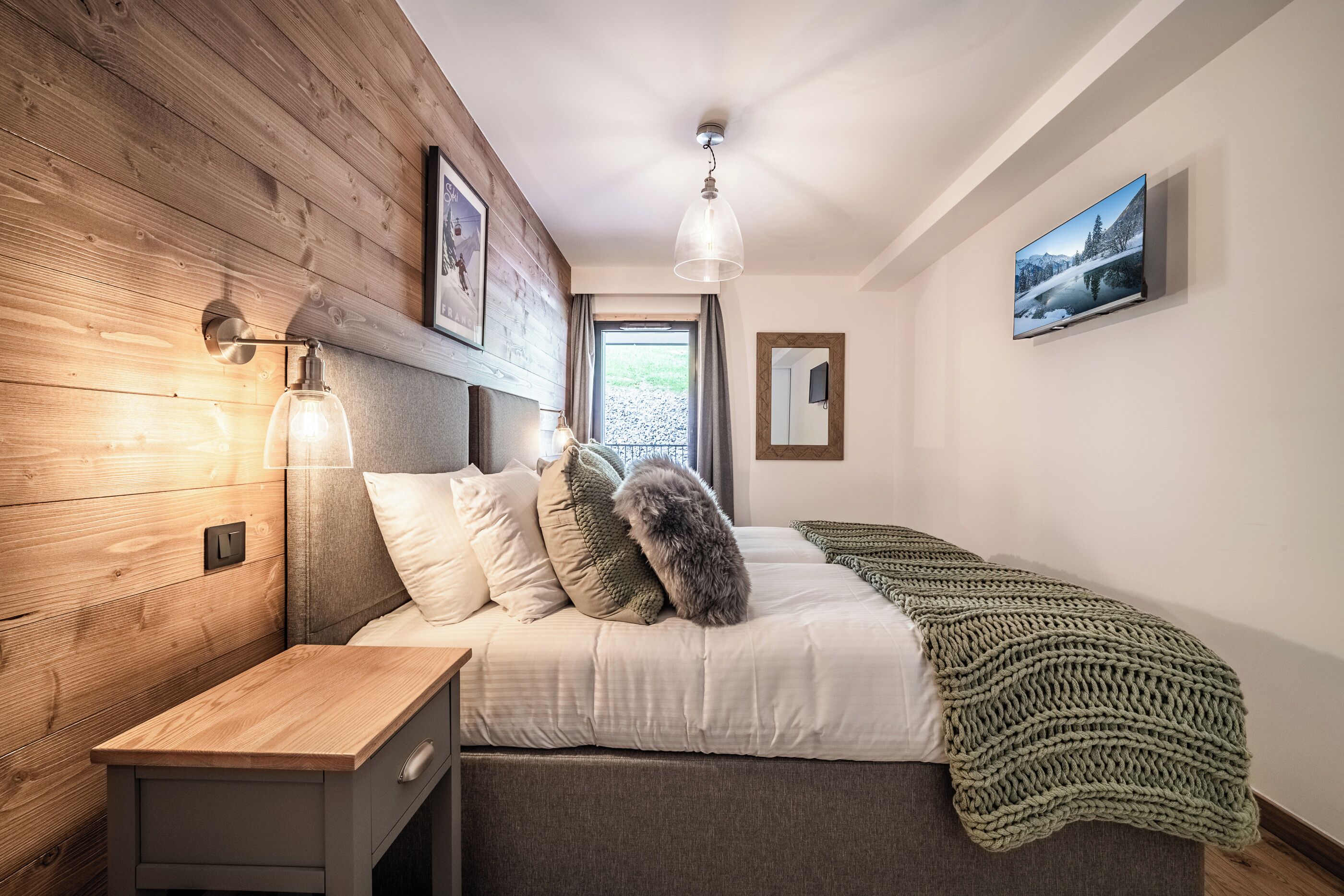 Un dormitorio con cabecera de madera, cama con manta verde, mesita de noche, televisor montado en la pared y ventana con vista a árboles.