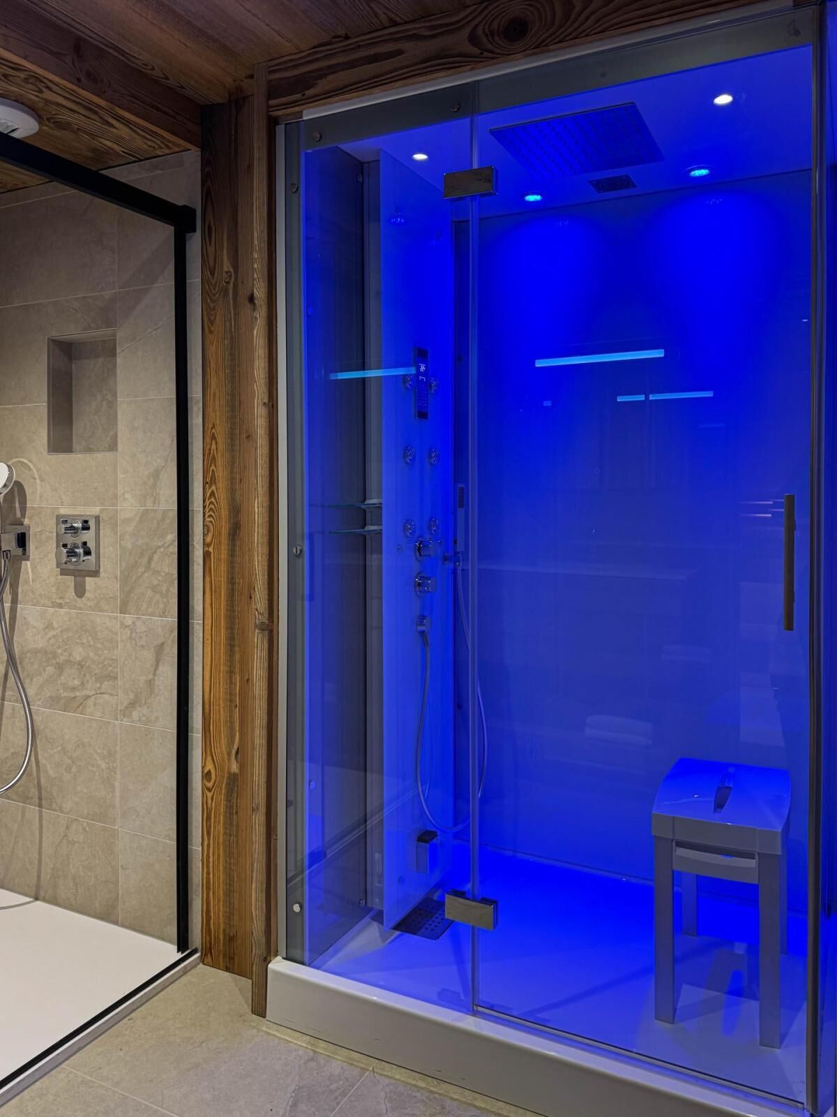 A modern shower enclosure with a glass door, a wooden frame, and a small stool inside.