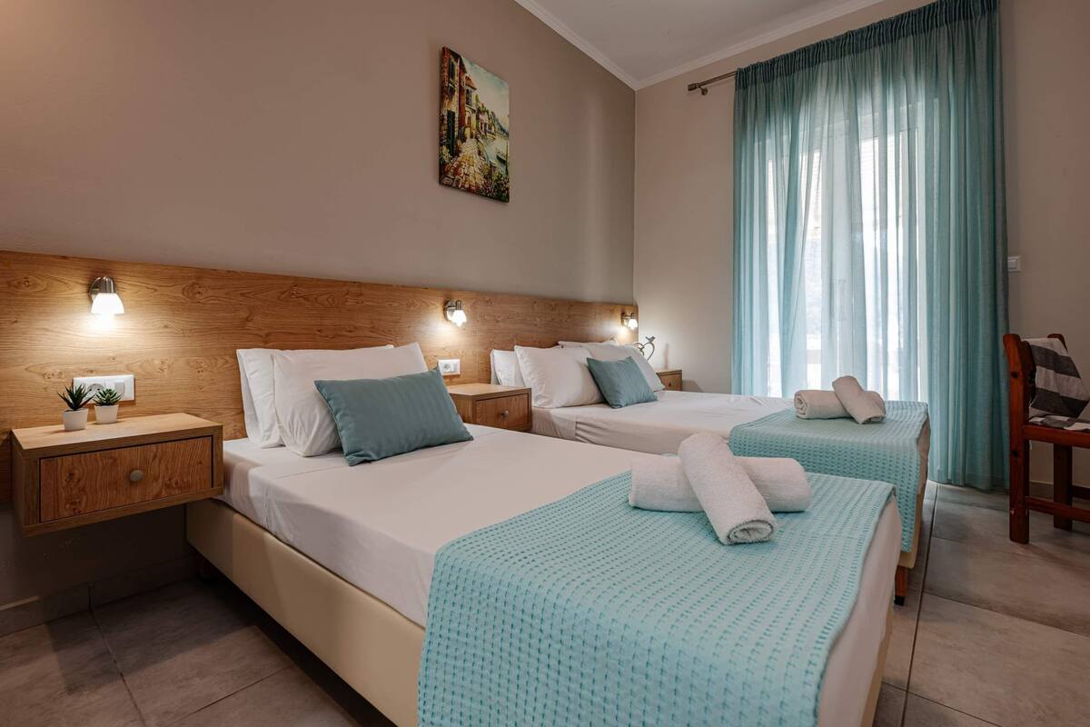 Une chambre d’hôtel avec deux lits, une tête de lit en bois, des lampes de chevet, un tableau au mur et une vue sur l’extérieur à travers des rideaux transparents.