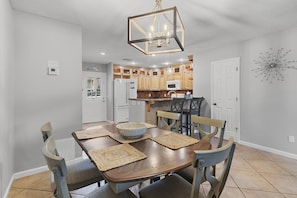 A dining area with a wooden table, chairs, a chandelier, and a kitchen in the background.