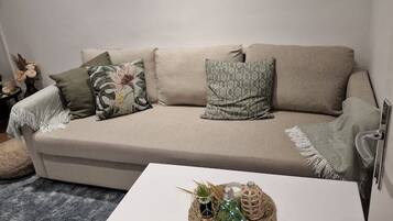 Een beige bank met gekleurde kussens, een geweven mandje op een witte tafel, en een vaas met gedroogde bloemen.
